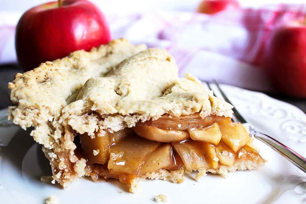 A slice of a gluten free vegan pie on a white plate with a fork and an apple behind it.