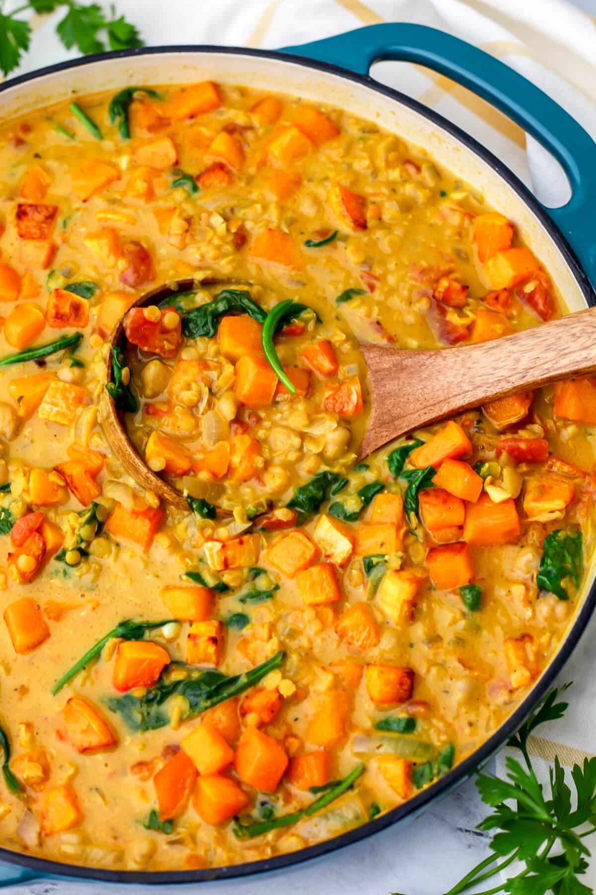 A large blue skillet full of Butternut squash and chickpea curry with red lentils and spinach with a wooden ladle in it.