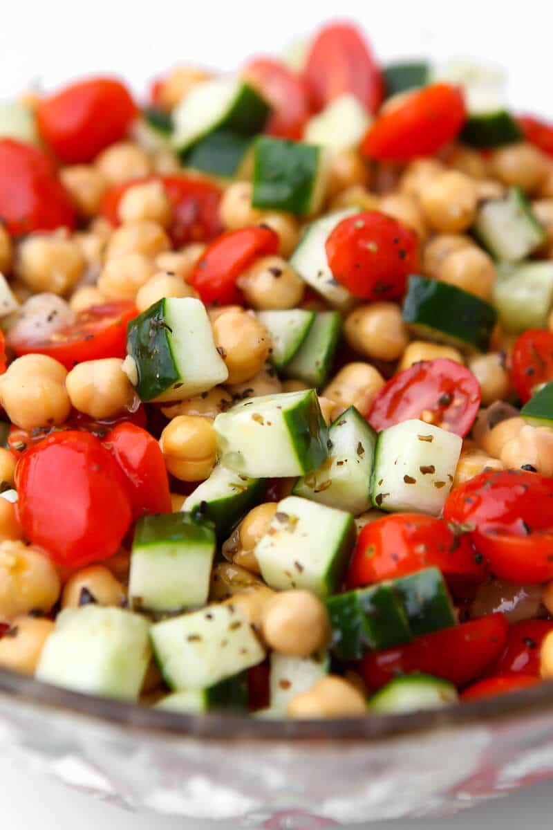 A glass bowl filled with chickpea salad with tomatoes and cucumbers with garlic vinaigrette dressing poured over it.