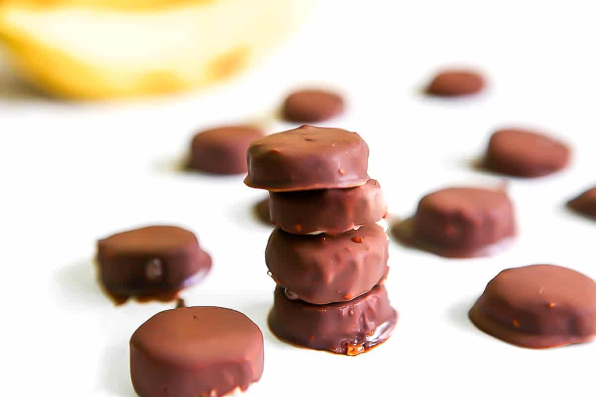 An assortment of vegan chocolate covered frozen banana bites on white parchment paper with a banana behind them.