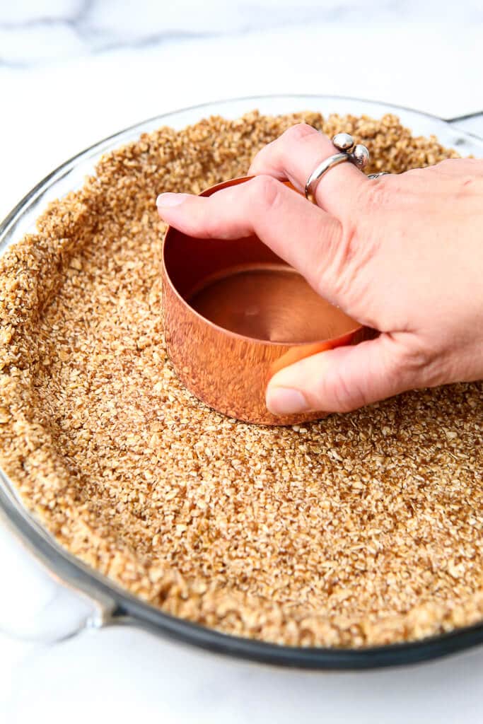 A gluten-free vegan graham cracker crust being packed down with the bottom of a copper measuring cup.