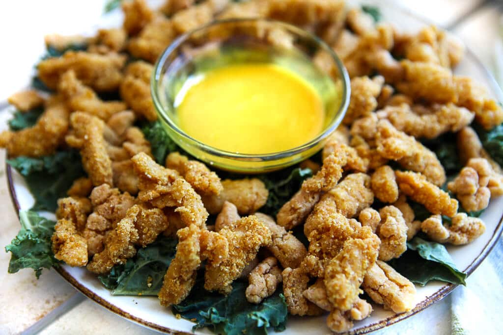 A plate full of vegan fried chicken strips on a bed of kale with sweet mustard dipping sauce. 