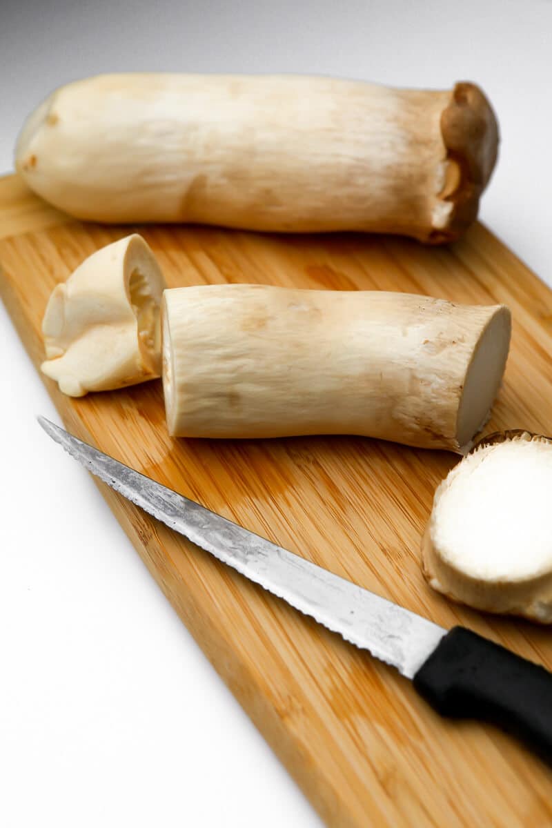 Two king oyster mushrooms on a cutting board with the top and bottom of the mushroom cut off of one of them.