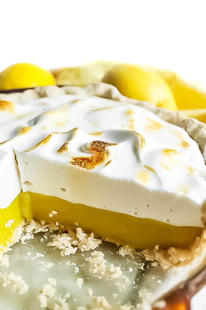 A pie dish with a vegan lemon meringue pie with a large slice taken out of it. Made with vegan lemon curd and aquafaba meringue topping.