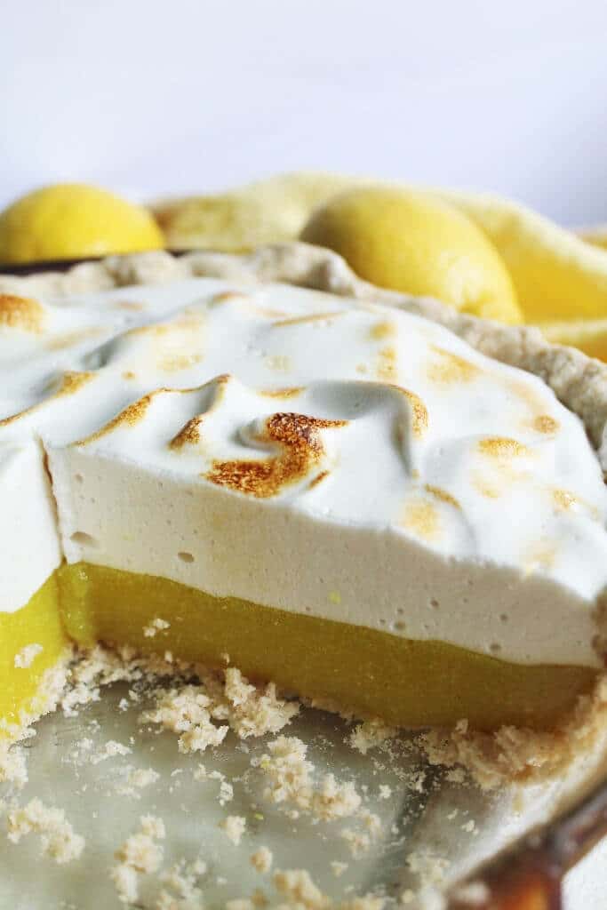 A pie dish with a vegan lemon meringue pie with a large slice taken out of it. Made with vegan lemon curd and aquafaba meringue topping.