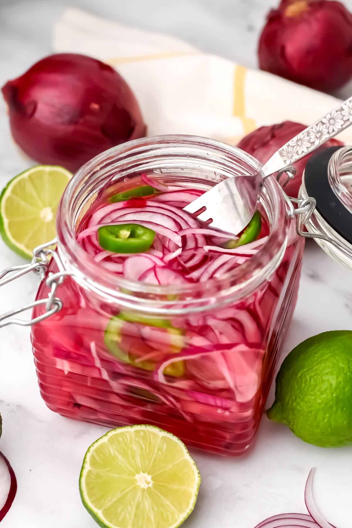 A glass jar filled with Mexican pickled onions with limes and red onions around it.