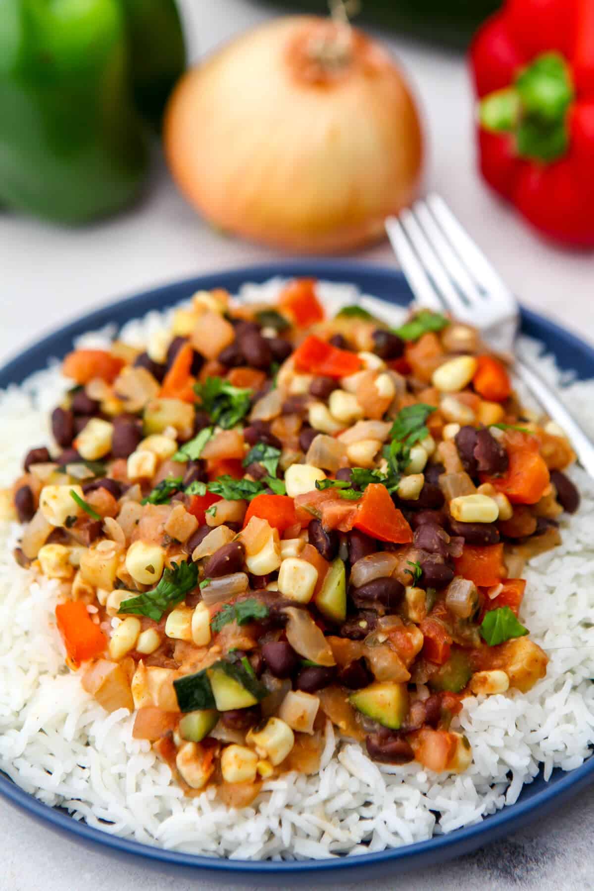 A blue plate filled with white rice and topped with a Mexican vegetable stir fry with veggies behind it.