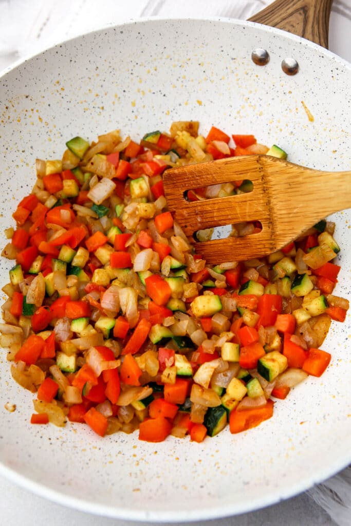 Diced onions, zucchini, and red bell peppers sautéing in a white skillet.