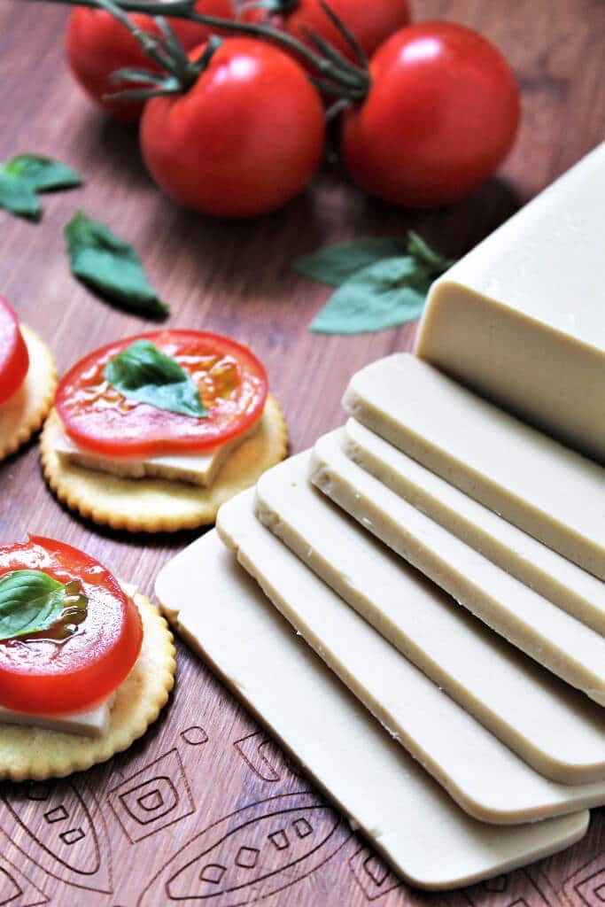 A block of vegan cheese sliced on a cutting board with tomatoes behind it.