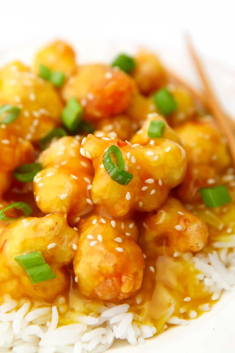 A plate of crispy fried cauliflower on a bed of rice with vegan orange sauce drizzled over top and chop sticks on the side.