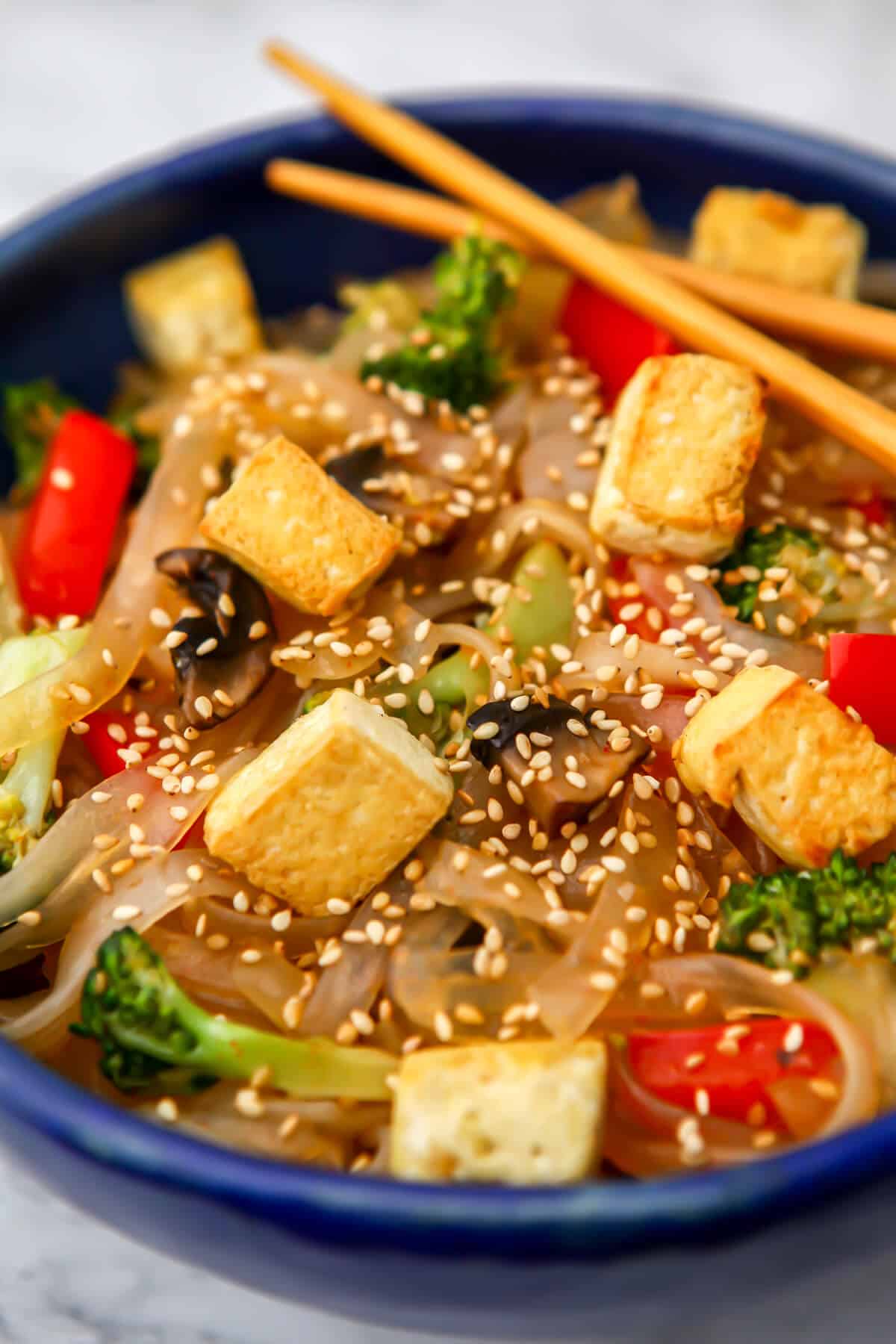 A blue bowl filled with spicy Korean glass noodles with stir fried veggies and tofu.