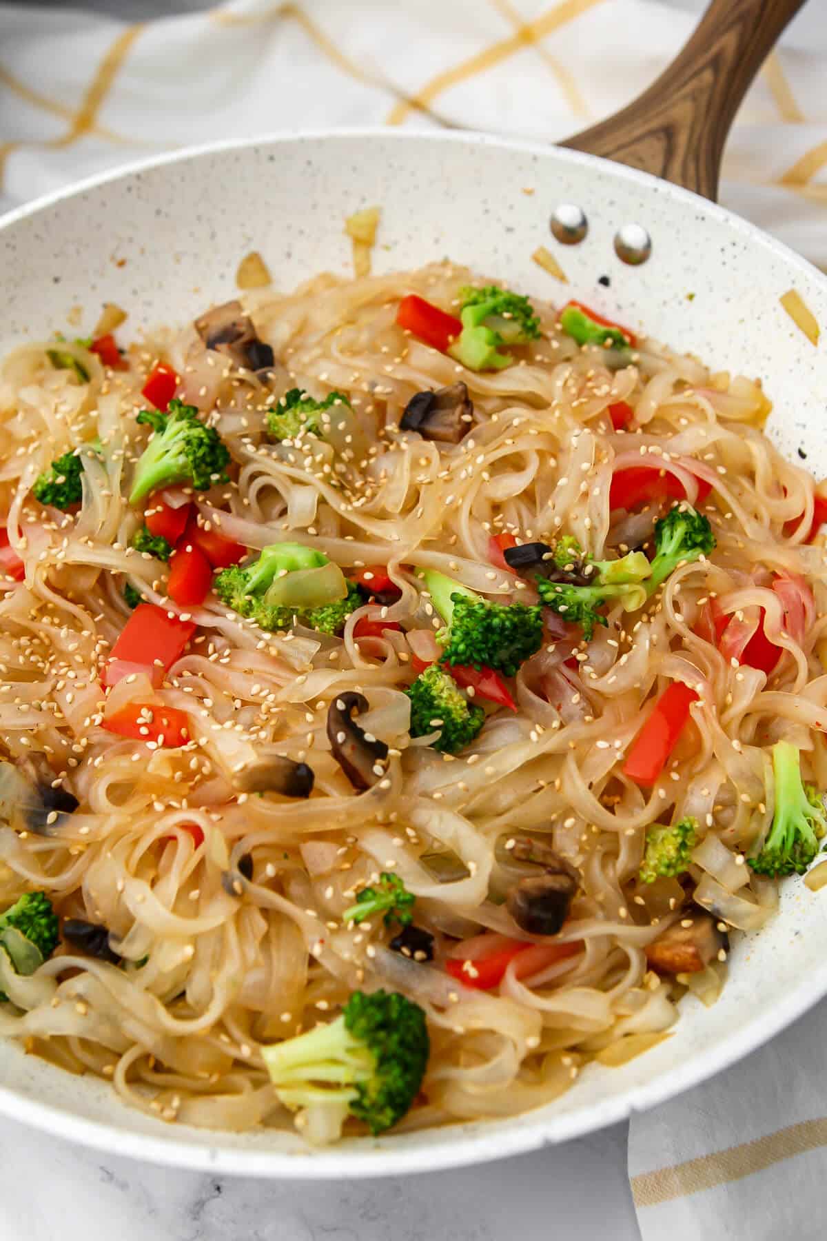 A white wok filled with glass noodle stir fry with a yellow and white tea towel behind it.