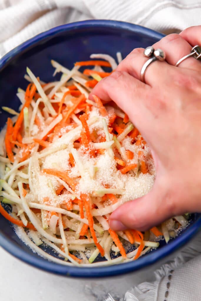 Shredded carrots and daikon in a blue bowl with salt and sugar sprinkled over them to make Bahn Mi pickled veggies.
