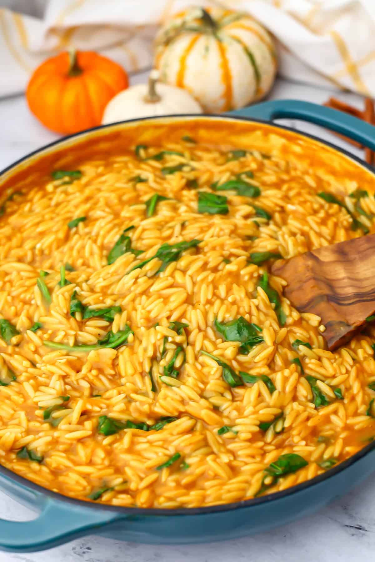 A large blue skillet with vegan  pumpkin orzo with spinach being stirred with a wooden spoon.