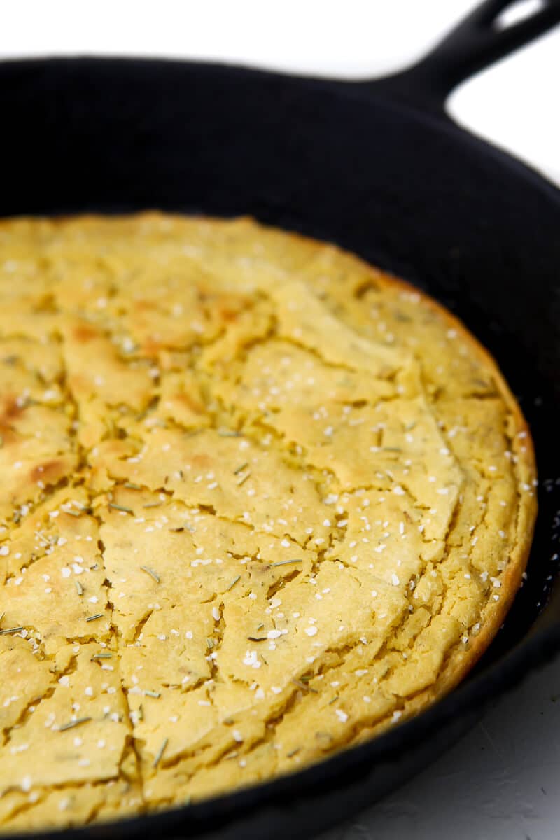 A close up of chickpea flatbread cooked in an iron skillet, sprinkled with salt and rosemary, and cut into wedges.