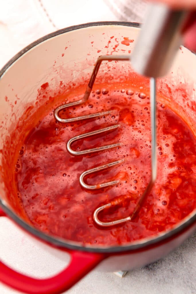 Cooked strawberries in a red saucepan being mashed with a potato masher.