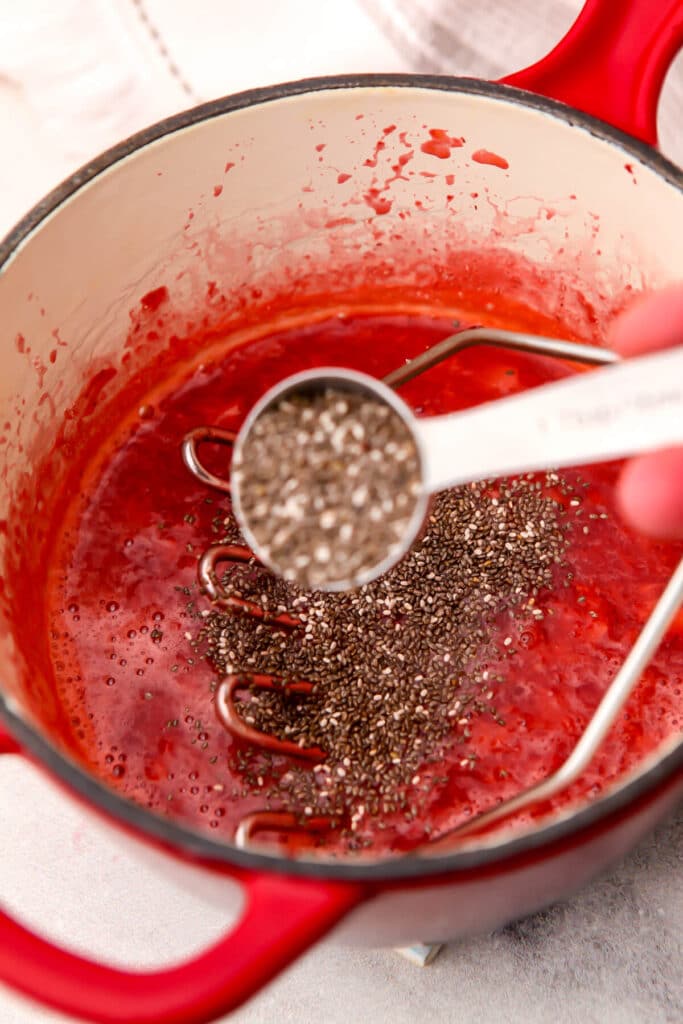 Chia seeds being added to a red sauce pan full of cooked strawberries to make chia jam.