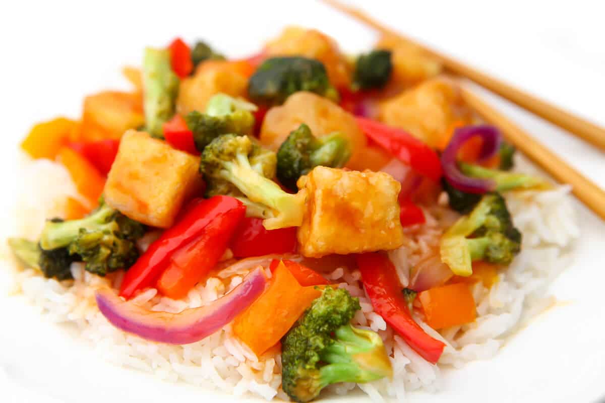 Veggies and tofu with sweet and sour sauce over white rice with chop sticks on the side.