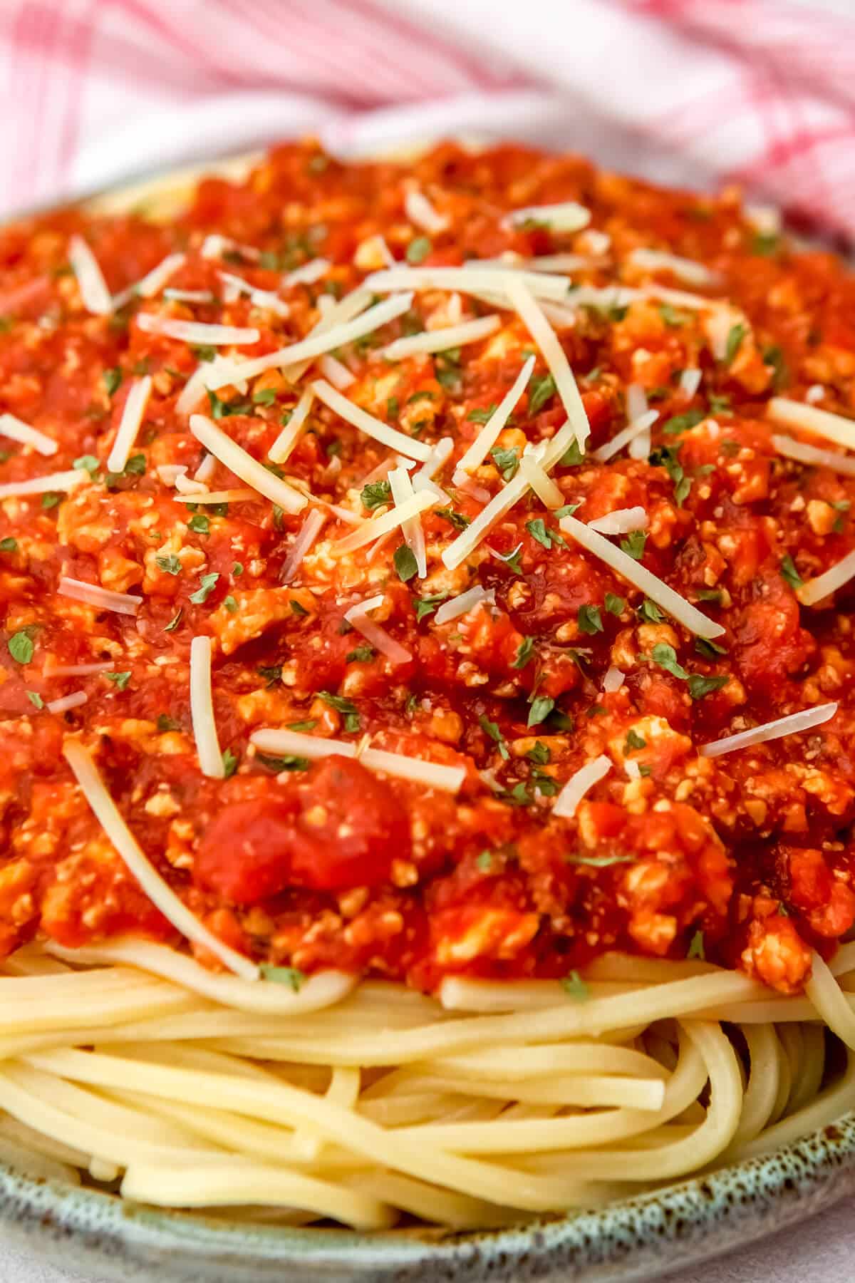 Meat like Bolognese sauce made from tofu served over pasta with a red and white tea towel behind the plate.