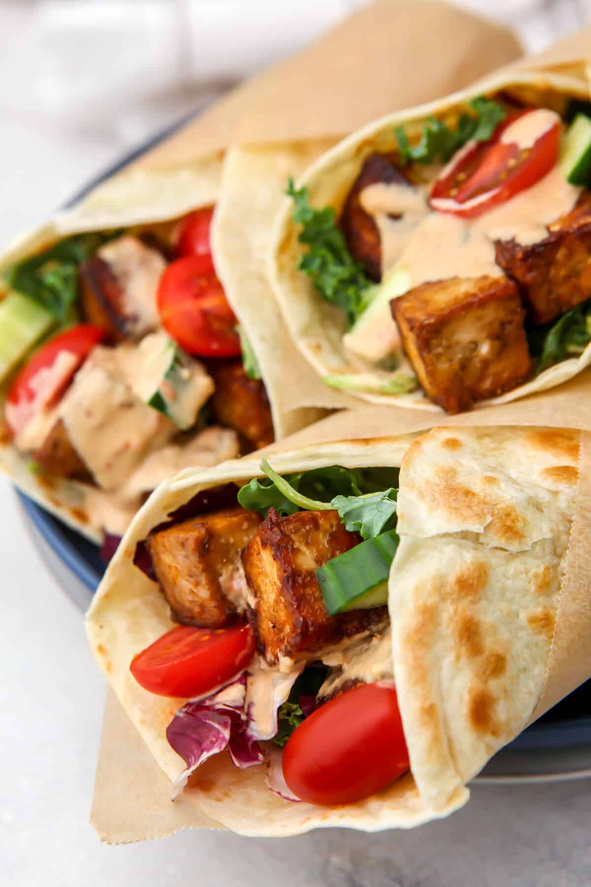 Three marinated tofu wraps with spicy tahini sauce and veggies stacked on a plate.