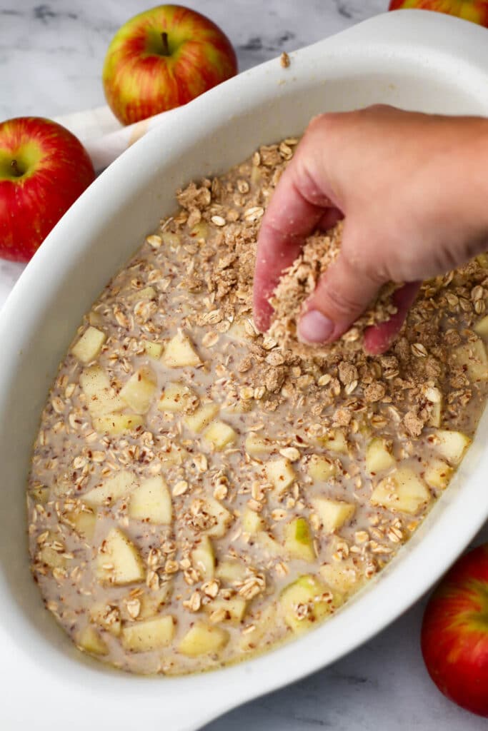 Baked oatmeal batter in a casserole dish being sprinkled with crumble topping before baking.