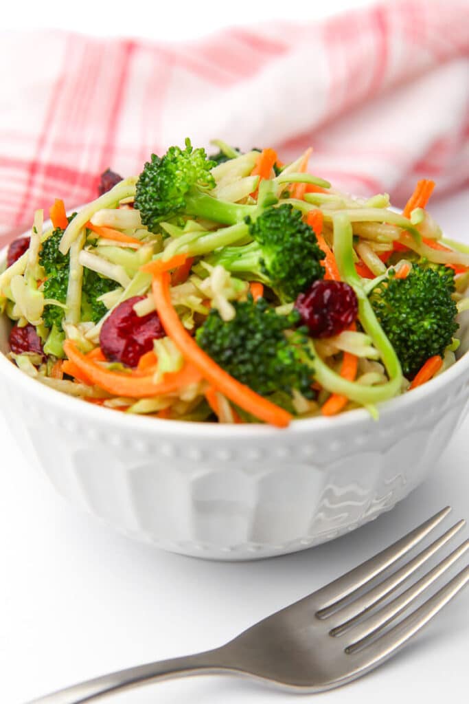 A white bowl filled with broccoli slaw salad with a fork in front and a red tea towel behind it.