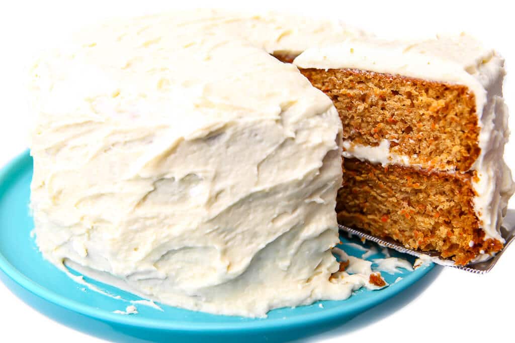 A piece of vegan carrot cake being taken out of a full cake on a blue cake stand.