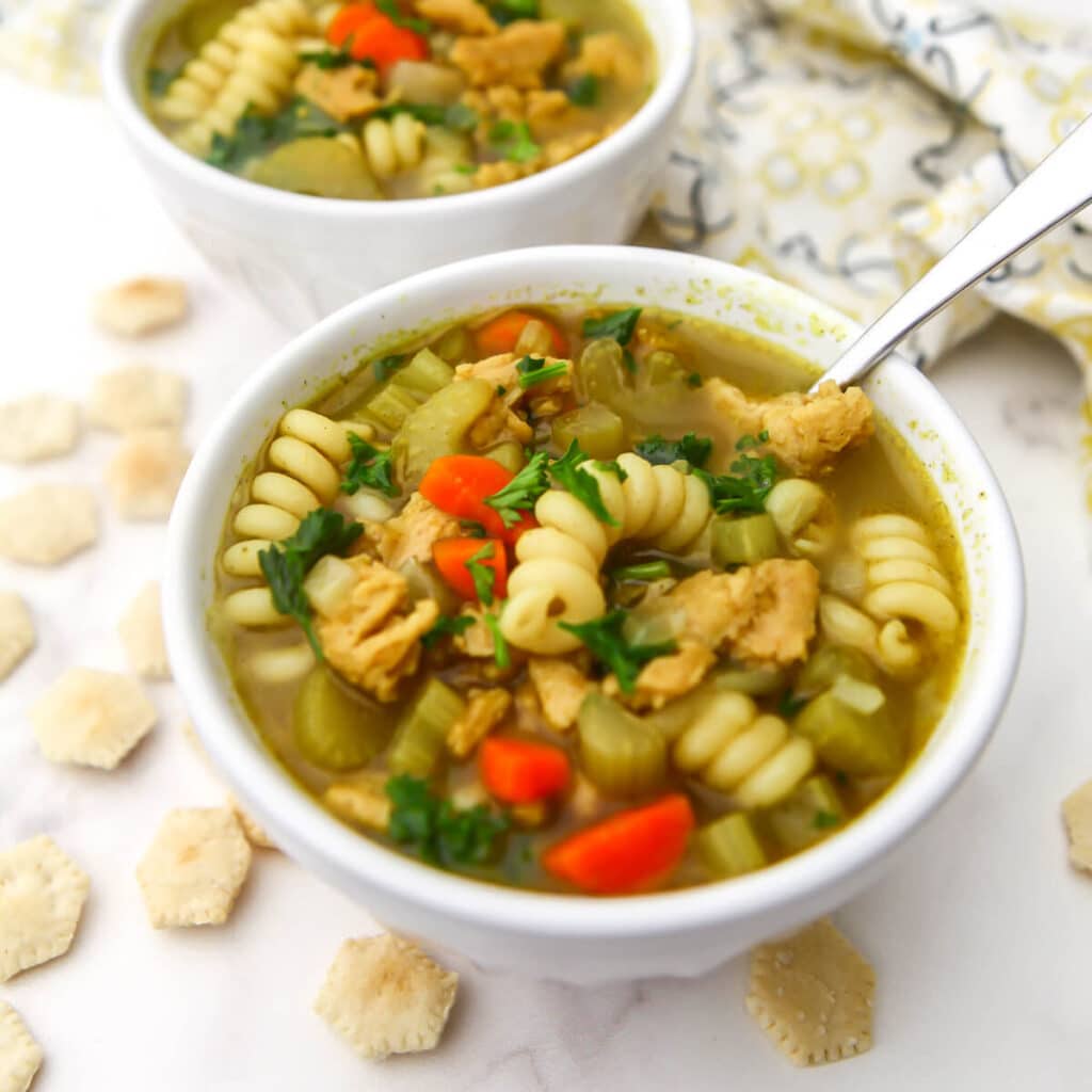 Two bowls of vegan chicken noodle soup made with soy curls with soup crackers around them.