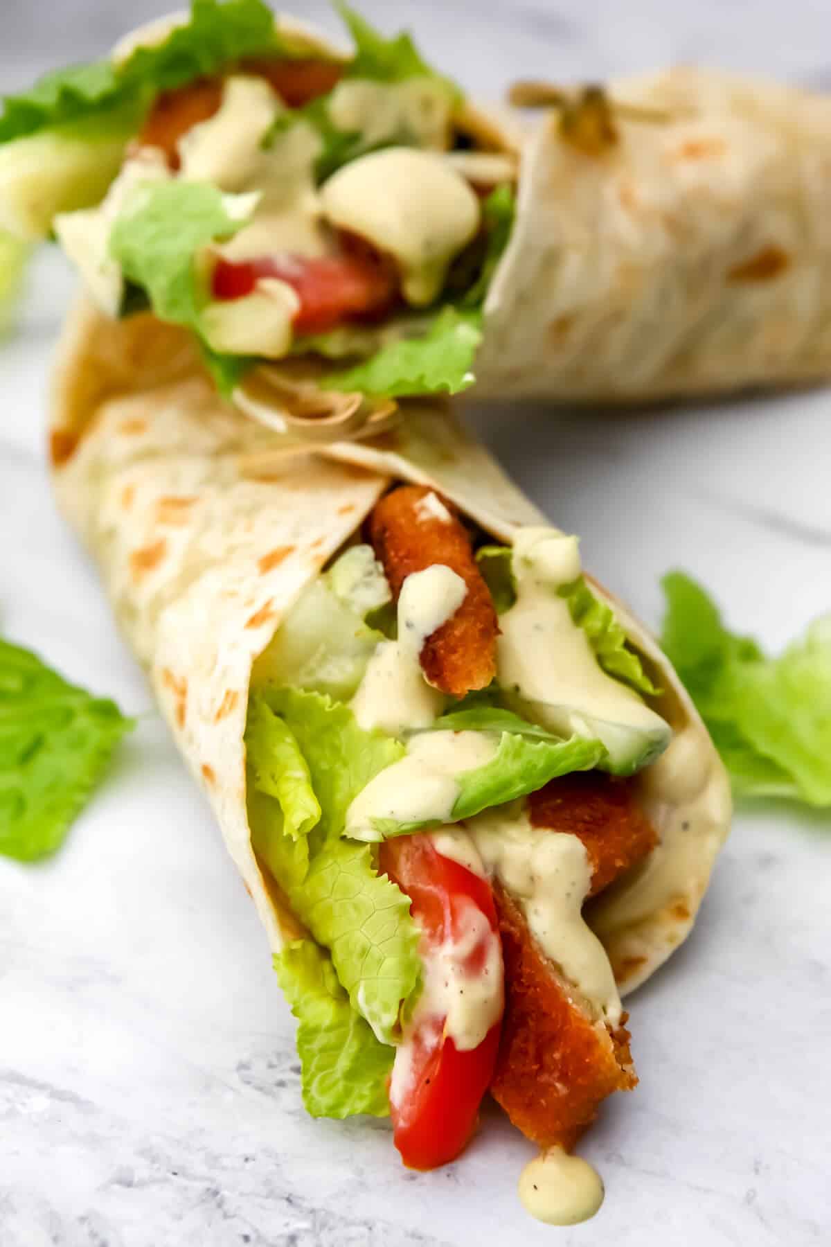 Two vegan chicken wraps with vegan Caesar, lettuce and veggies on a marble countertop.