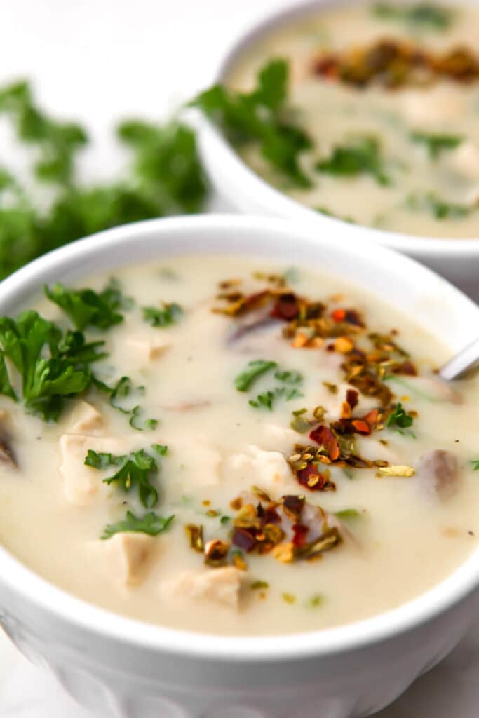 Two white bowls filled with vegan cream of chicken soup with parsley and red and green pepper flakes on top.