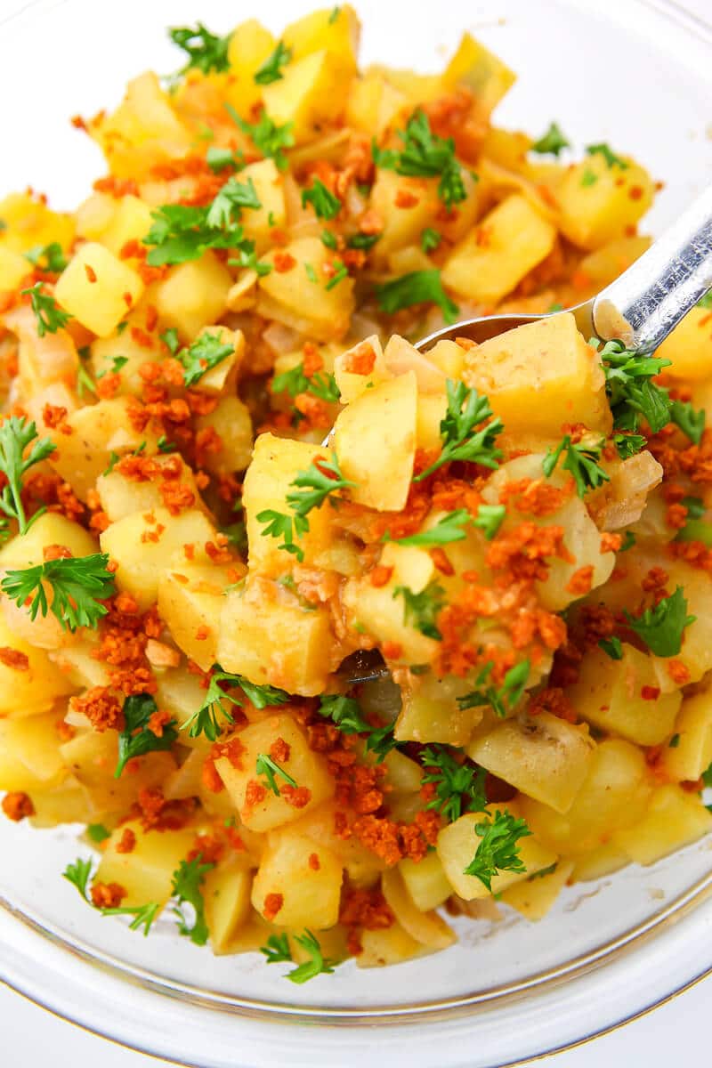 A top view of a German potato salad made with vegan bacon bits being scooped out of a bowl.