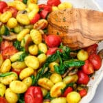 Vegan gnocchi with tomaotes, mushrooms, onions, and spinch frying in a pan being stirred with a wooden spoon.