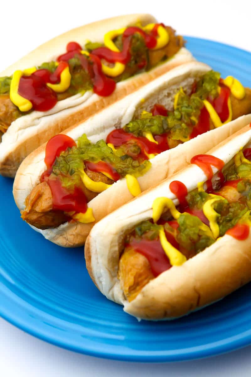 Three vegan hot dogs with the works on them sitting on a blue plate.