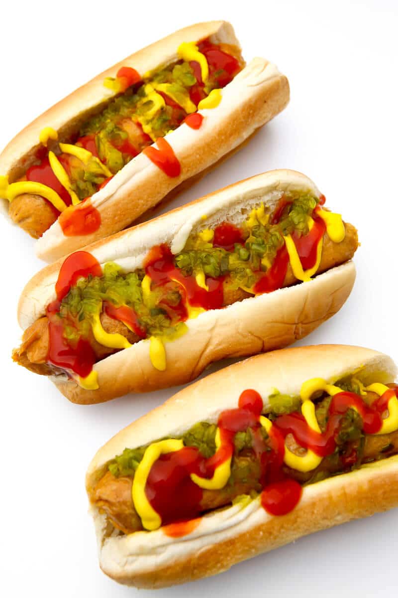 Three vegetarian hot dogs made from seitan in buns with mustard, ketchup, and relish on them on a white background.