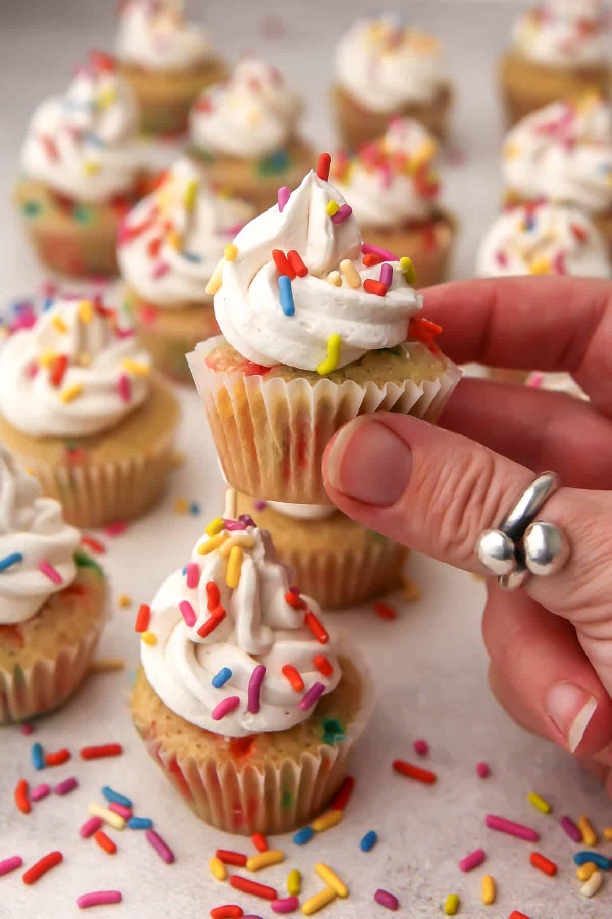 Vegan funfetti mini cupcakes with sprinkles on a countertop with someone holding up one.