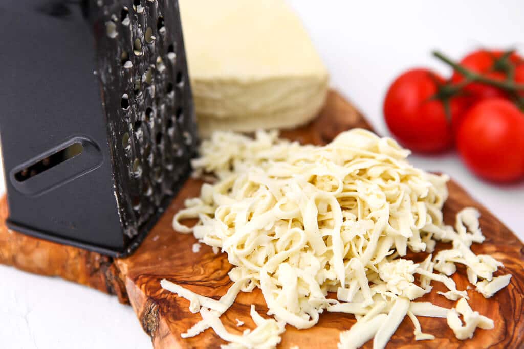 Dairy-free mozzarella cheese on a cutting board with a cheese grater and a tomato on the side.