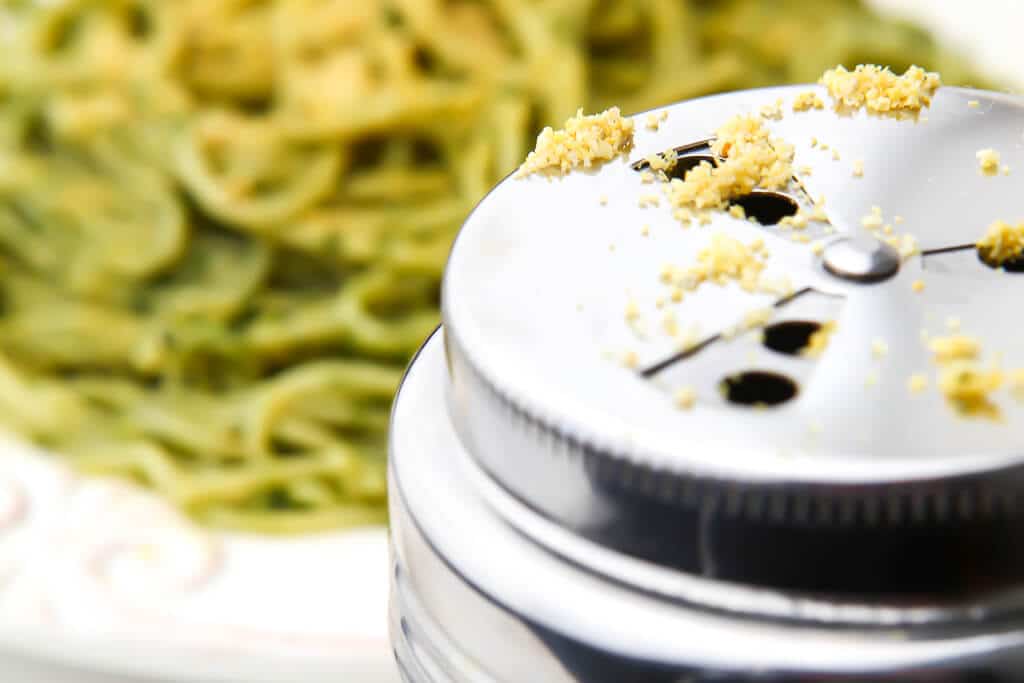 A silver Parmesan cheese shaker filled with vegan Parmesan in front of a plate of spaghetti with pesto.