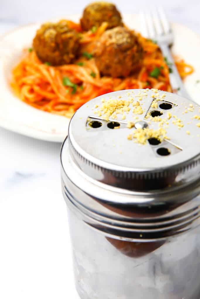 A Parmesan cheese shaker filled with vegan Parmesan in front of a plate of spaghetti and meatballs.