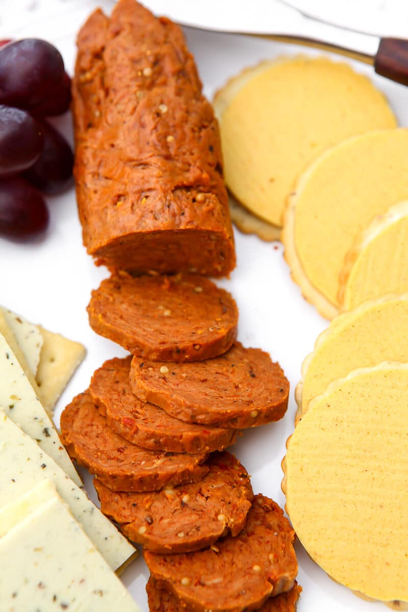 A stick of veggie pepperoni on a white plate with vegan cheese and grapes on the side.