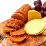 Homemade vegan pepperoni on a wooden cutting board next to sliced cheese and purple grapes.