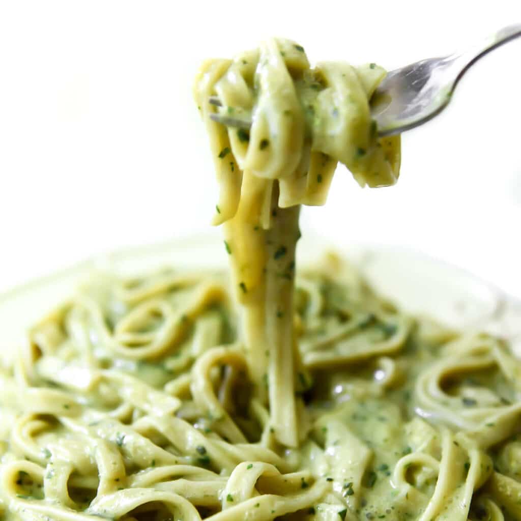 A plate filled with creamy vegan pesto sauce over pasta and a fork full of the pasta over the dish.