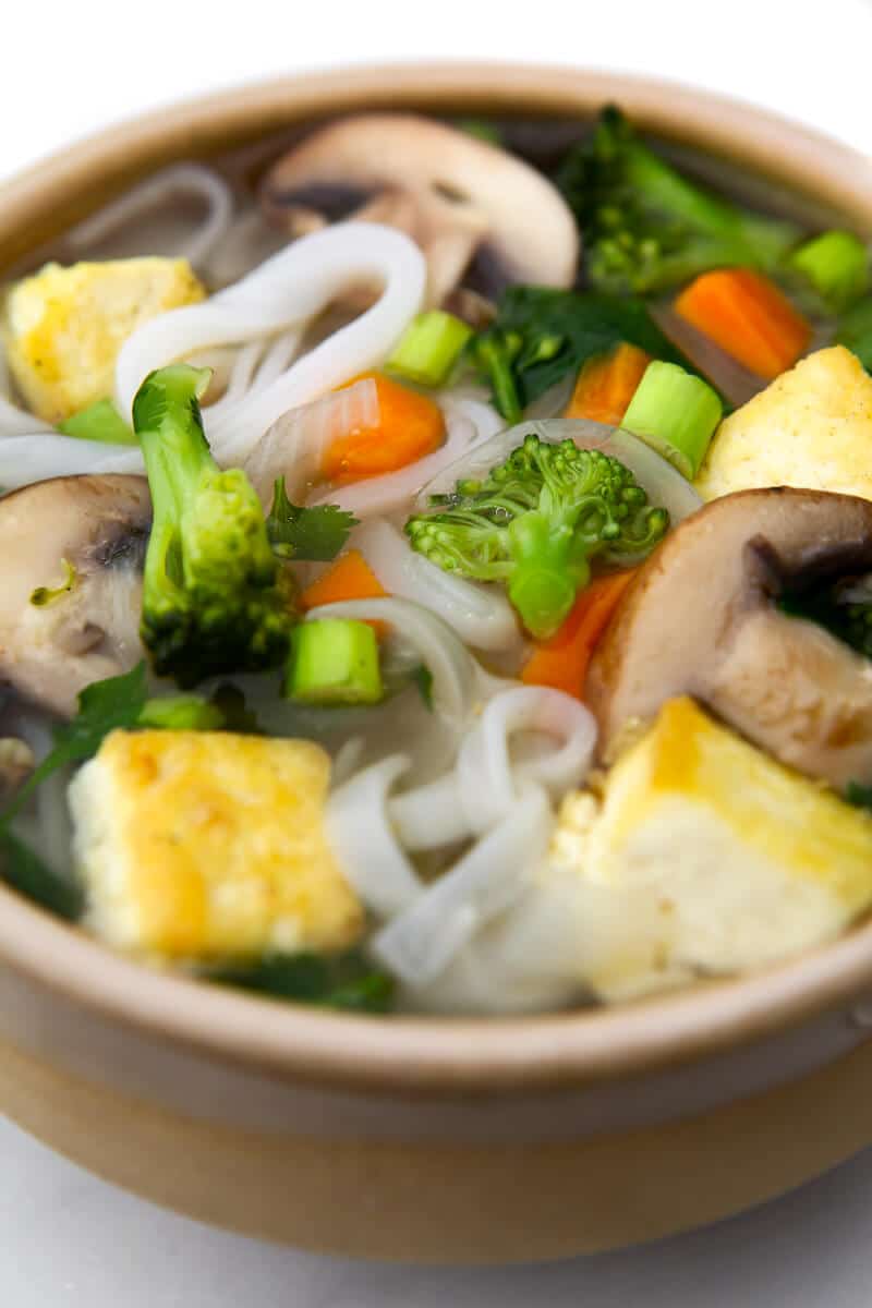 A brown bowl filled with rice noodles, tofu, veggies, and pho soup broth.