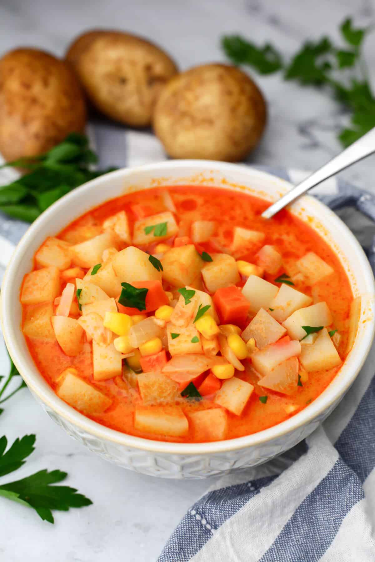 A bowl filled with vegan potato corn chowder in a creamy smoked paprika broth. with potatoes in the background and a blue and white tea towel.