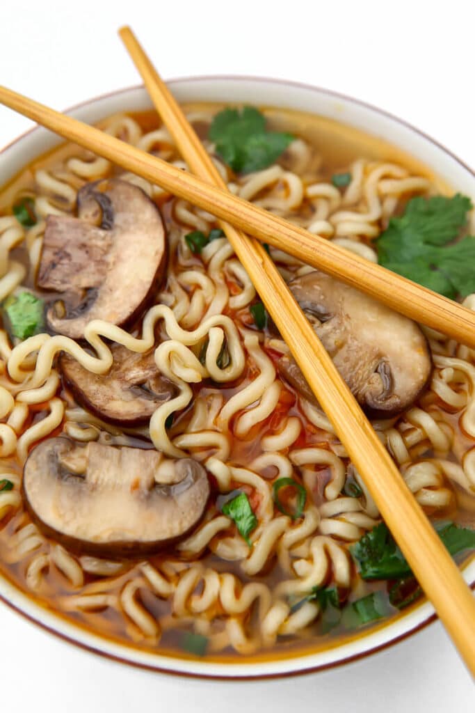 A bowl of ramen with mushrooms, cilantro, and green onions with chop sticks resting on top of the bowl.