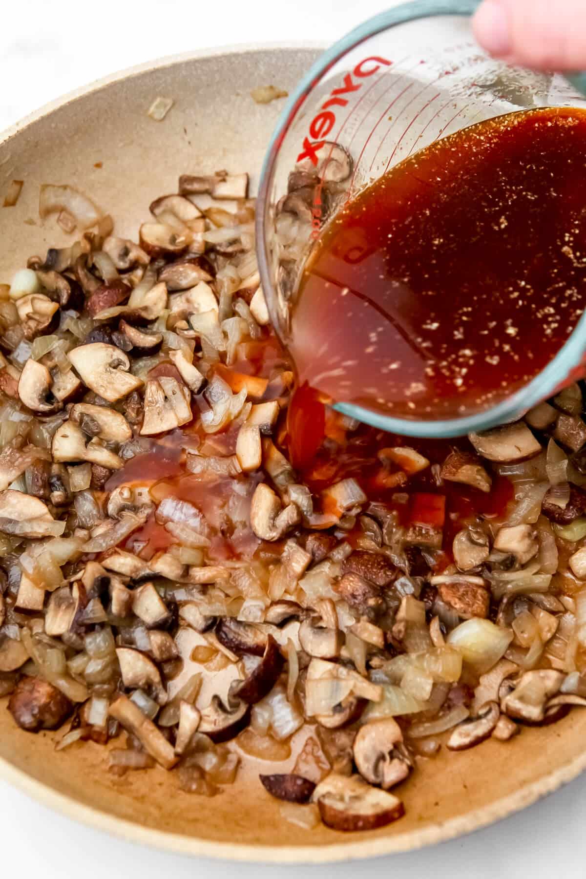 Broth being added to sauteed onions and mushrooms in a frying pan.