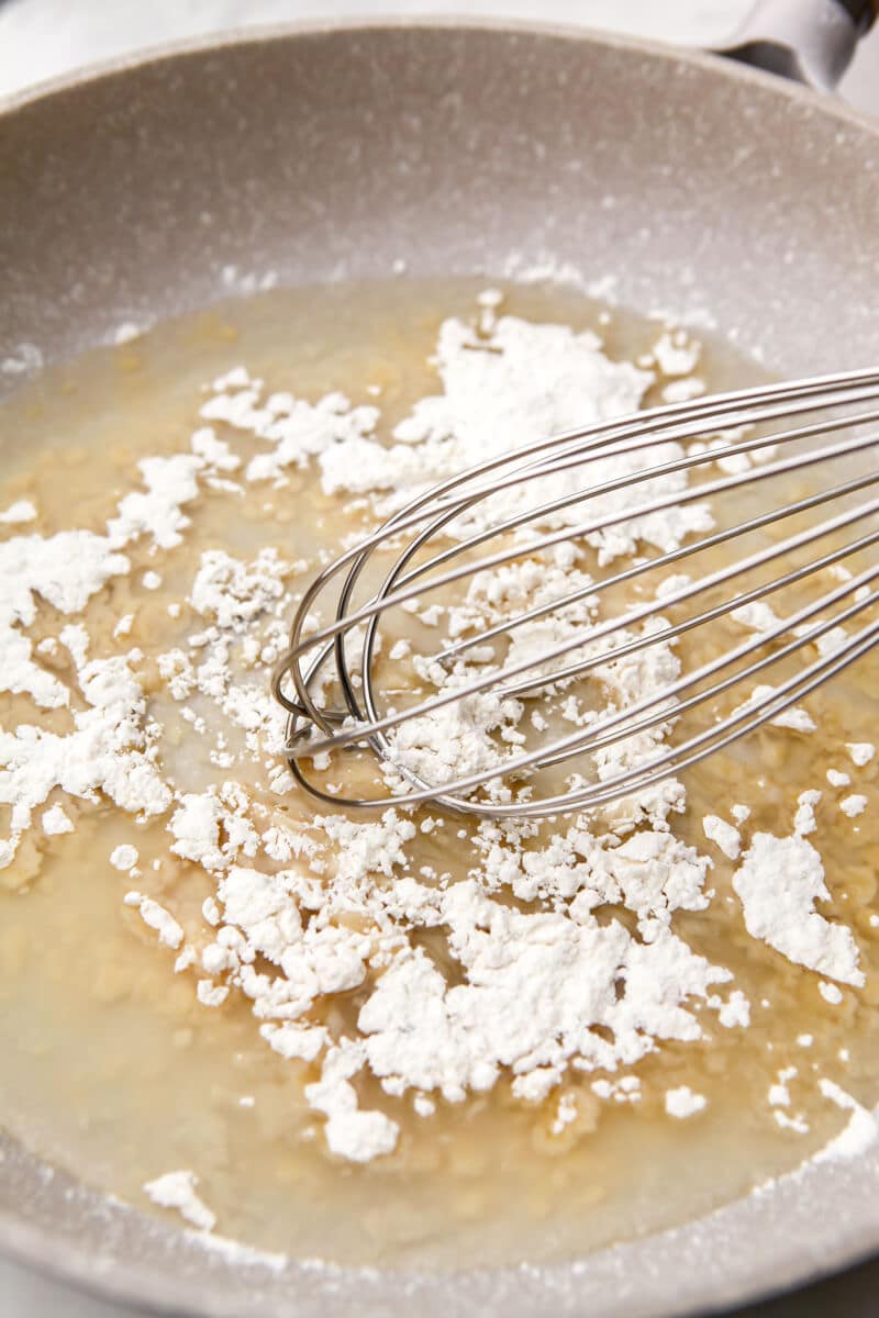 A frying pan with oil and flour being stirred with a whisk to make a roux.