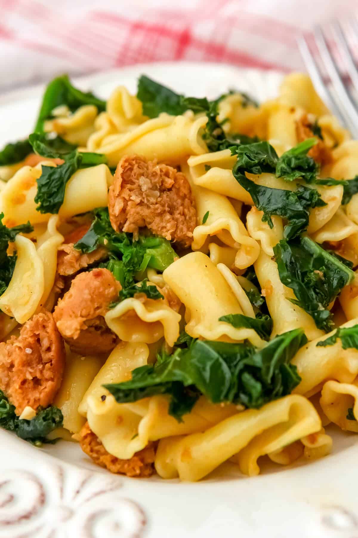 Vegan sausage and kale pasta on a white plate with a red tea towel behind it.