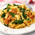 Vegan sausage pasta with kale on a white plate with a fork on the side.