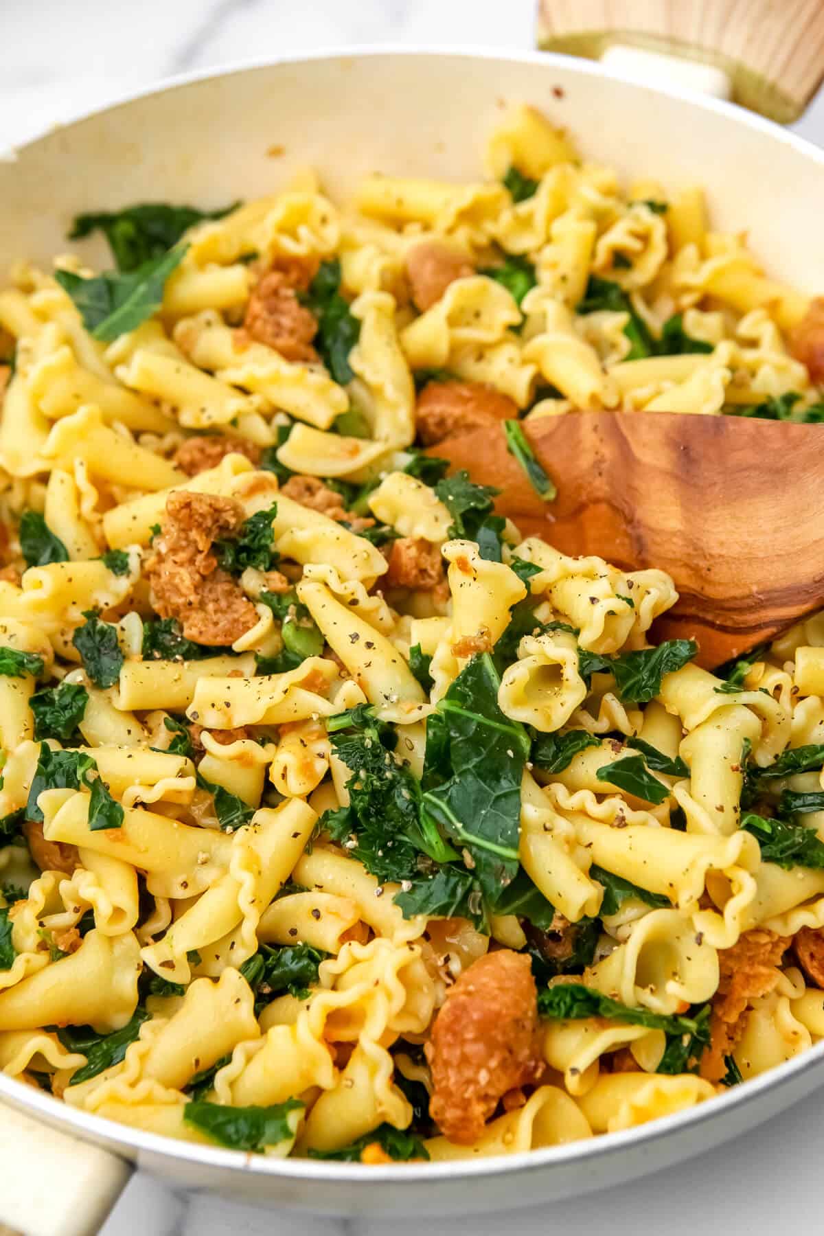 A large pot filled with noodles, sausage and kale being stirred with a wooden spoon.