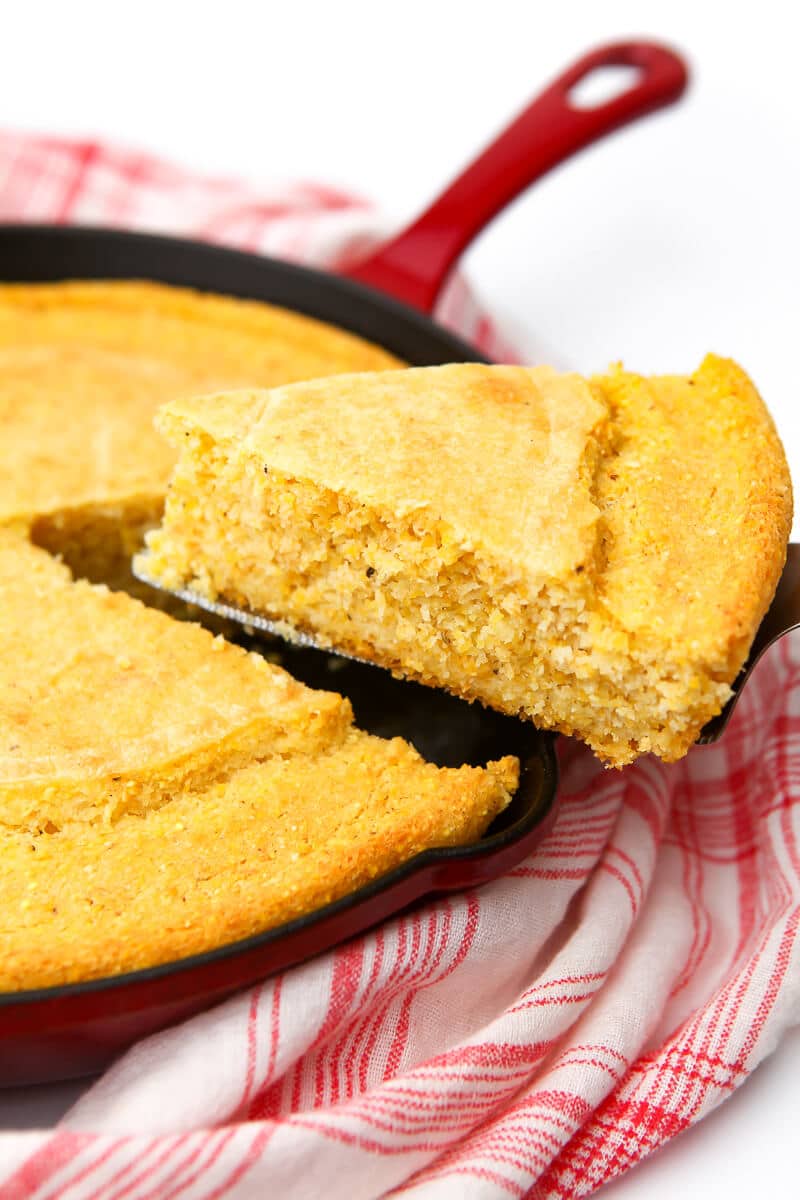 A slice of vegan skillet cornbread being taking out of a red skillet.